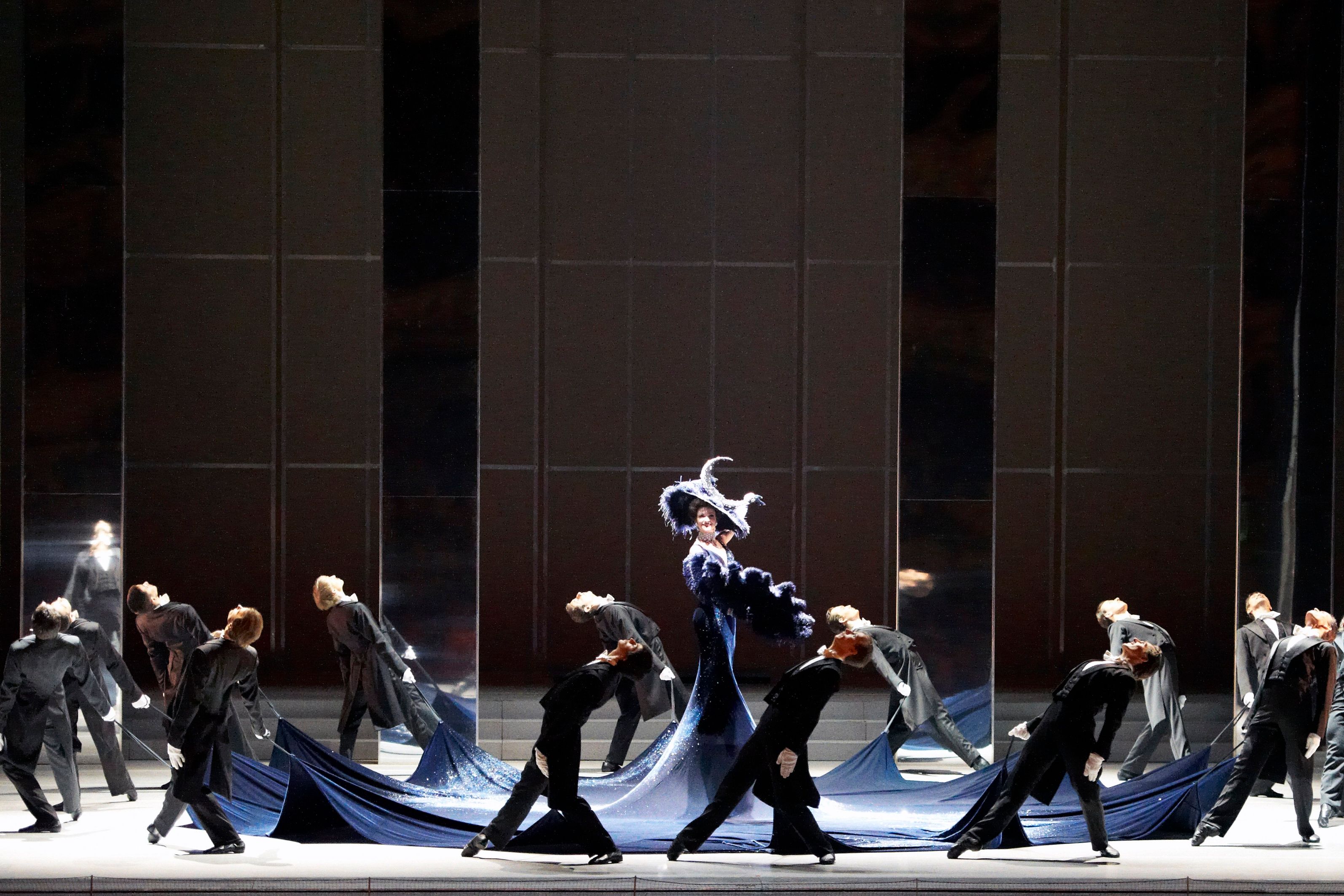 Scene photo from the ballet “Die Fledermaus” at the Vienna State Opera. Dance scene with dancers in black tailcoats dancing around a lady with a sweeping hat and fan by holding the ends of her dark blue, extremely long dress